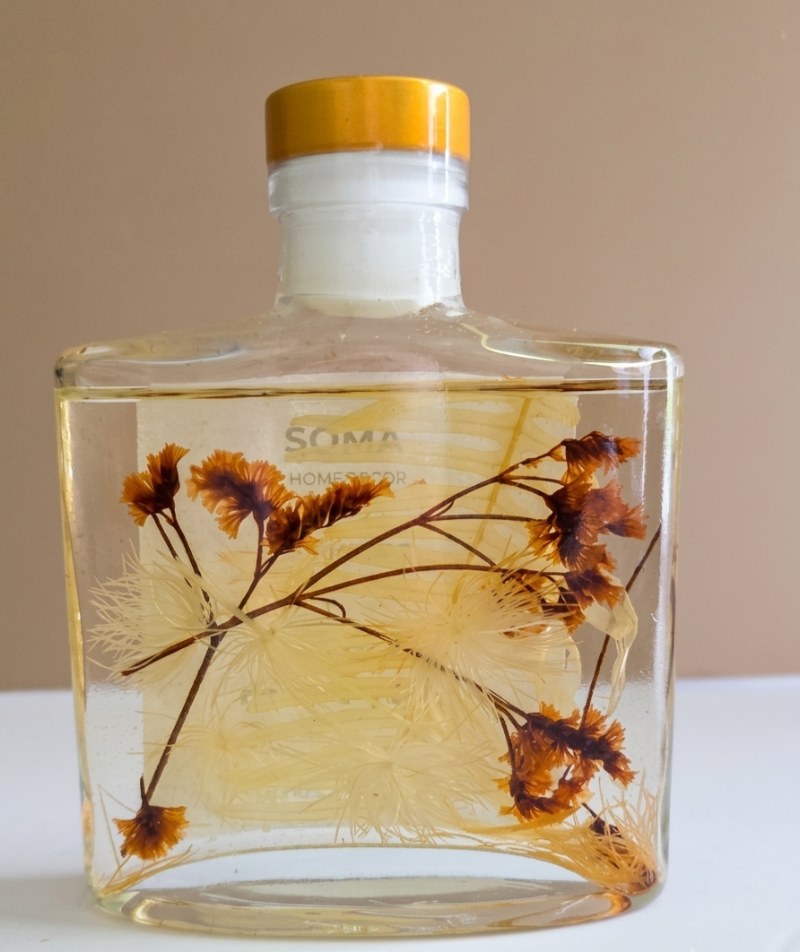A clear glass bottle with a gold cap containing a pale yellow liquid and dried botanical elements, including orange-brown flowers and fine thread-like strands. The word 'SOMA' is printed on the front, set against a plain beige background.