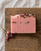 A rectangular bar of pink wild rose soap sits on a dark wooden board, with dried rose petals on top and a few loose rose buds nearby. Text below the soap lists ingredients and identifies the brand as SOMA HOME DECOR LLC.