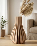 A stylish 3D-printed vase with a unique ribbed design and a warm, wooden texture, holding a bouquet of dried pampas grass and palm leaves on a wooden table in a brightly lit, modern living room. A small, elegant brand card sits next to it.