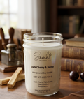 A handpoured soy candle in a clear glass jar with a white wax fill and a white printed label reading 'Soma Home' and 'Dark Cherry & Santo'. The candle sits on a wooden table in a warm, softly blurred room with books and wooden objects in the background.
