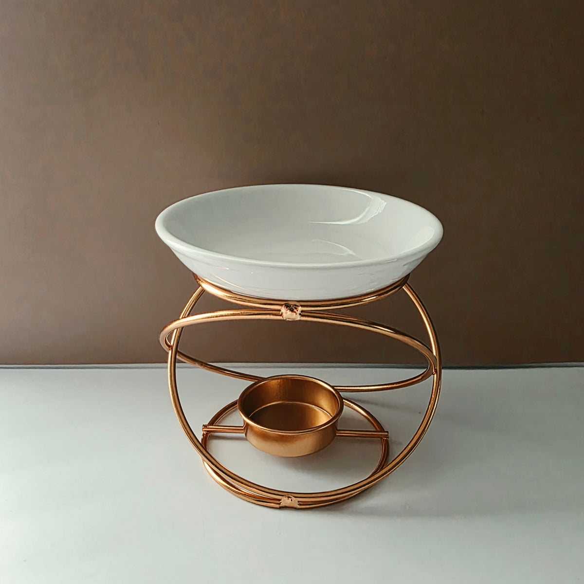 A white ceramic bowl displayed on a copper-toned metal stand with a circular base and a smaller matching bowl inside, set against a brown wall and light surface.