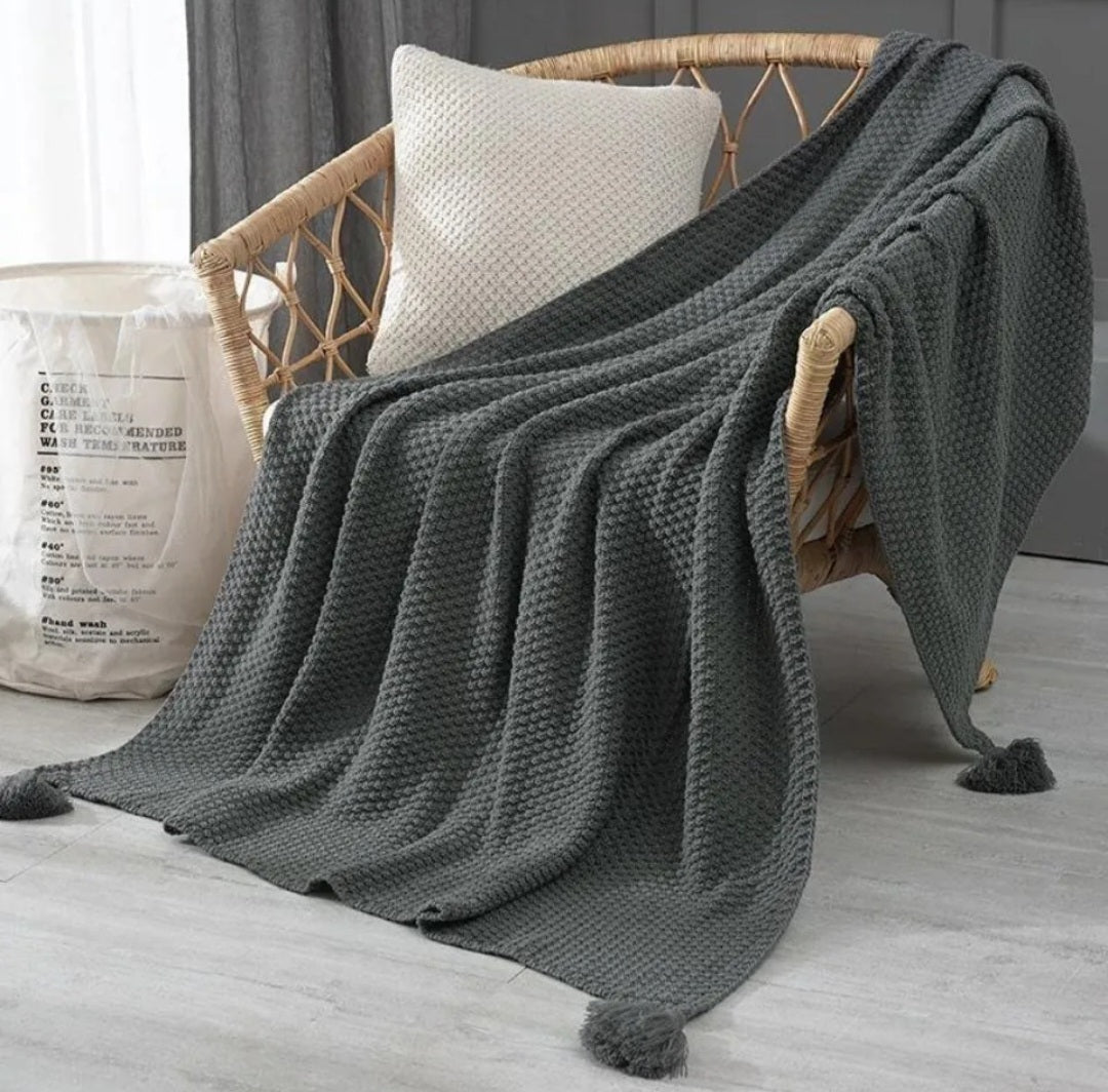 A dark gray waffle-weave throw blanket with pom-pom tassels draped over a rattan armchair with a white pillow, next to a white fabric storage bag on a light gray floor.