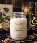 A handpoured soy candle in a clear glass mason jar by Somal Home, labeled 'Medusa's Elixir,' set on a rustic wooden surface with dried herbs, dark plums, and books in the background.