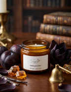 An amber glass jar candle labeled 'Somni Home - Medusa's Elixir' sits on a wooden table surrounded by plums, amber crystals, dried vanilla beans, and a small brass bell, with a blurred bookshelf in the background.