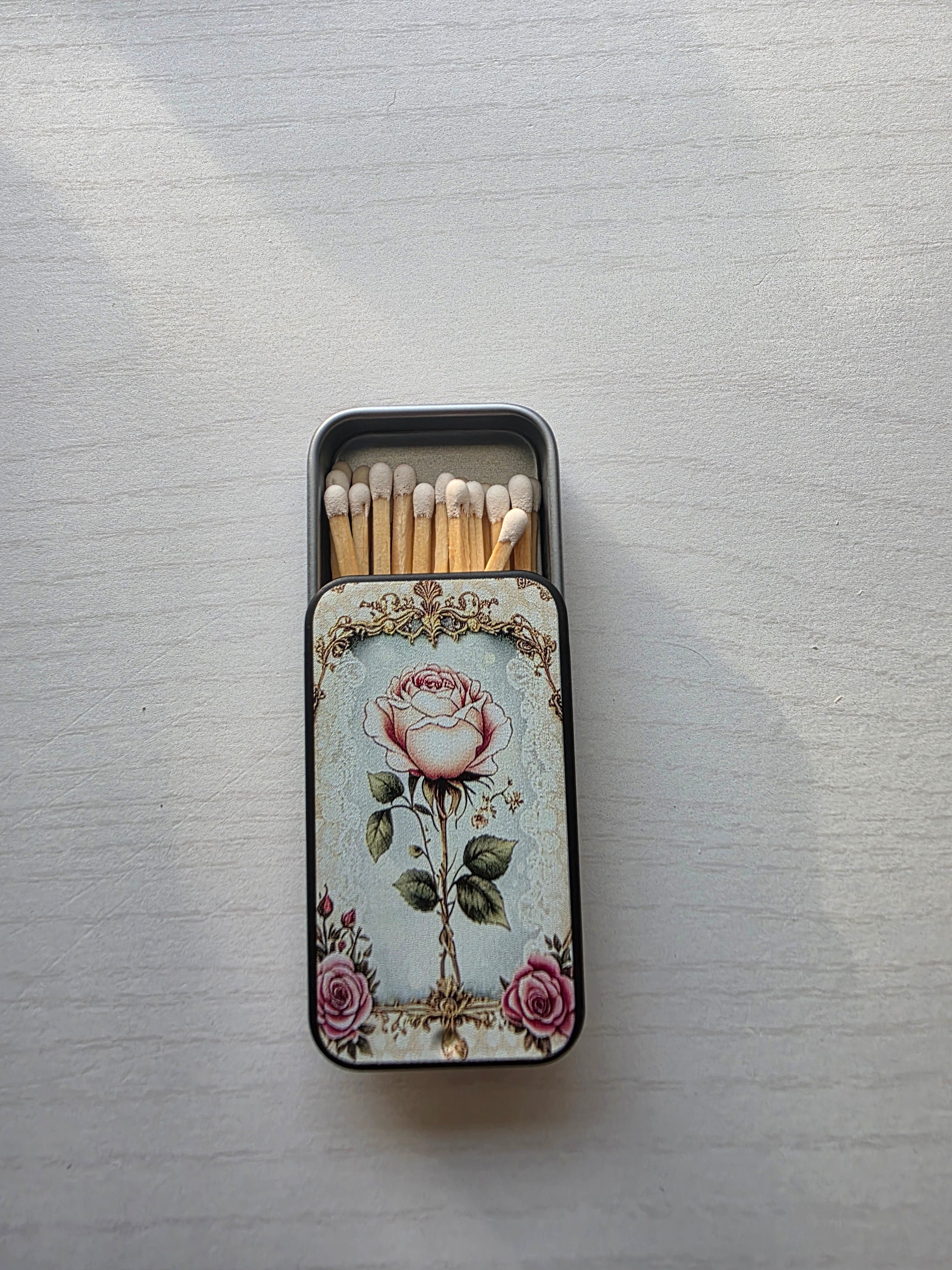 A decorative metal matchbox with a vintage-style pink rose illustration on the front, shown open with wooden matches inside on a light textured background.