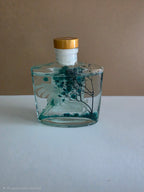 A clear glass bottle with a gold cap and white neck, containing a teal liquid with dried botanical elements, set against a two-tone beige and white background.