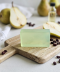 A translucent pale yellow-green rectangular bar of soap sits on a wooden cutting board, surrounded by sliced pears, dried berries, and a small bottle in a softly blurred kitchen-style setting.