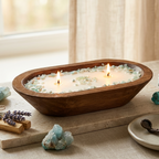 A wooden bowl-shaped candle holder with two lit candles, filled with white wax and decorated with light blue and green crystal chips, styled on a linen tablecloth with crystals and lavender.