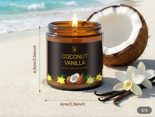 A lit amber glass candle jar labeled 'Coconut Vanilla' sits on a sandy beach with a half coconut, vanilla beans, and white flowers in the background. Measurement text showing 6.