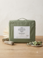 An olive green Melanni cotton sheet set package with a white label, styled on a wooden surface with dried flowers and greenery against a textured off-white wall.
