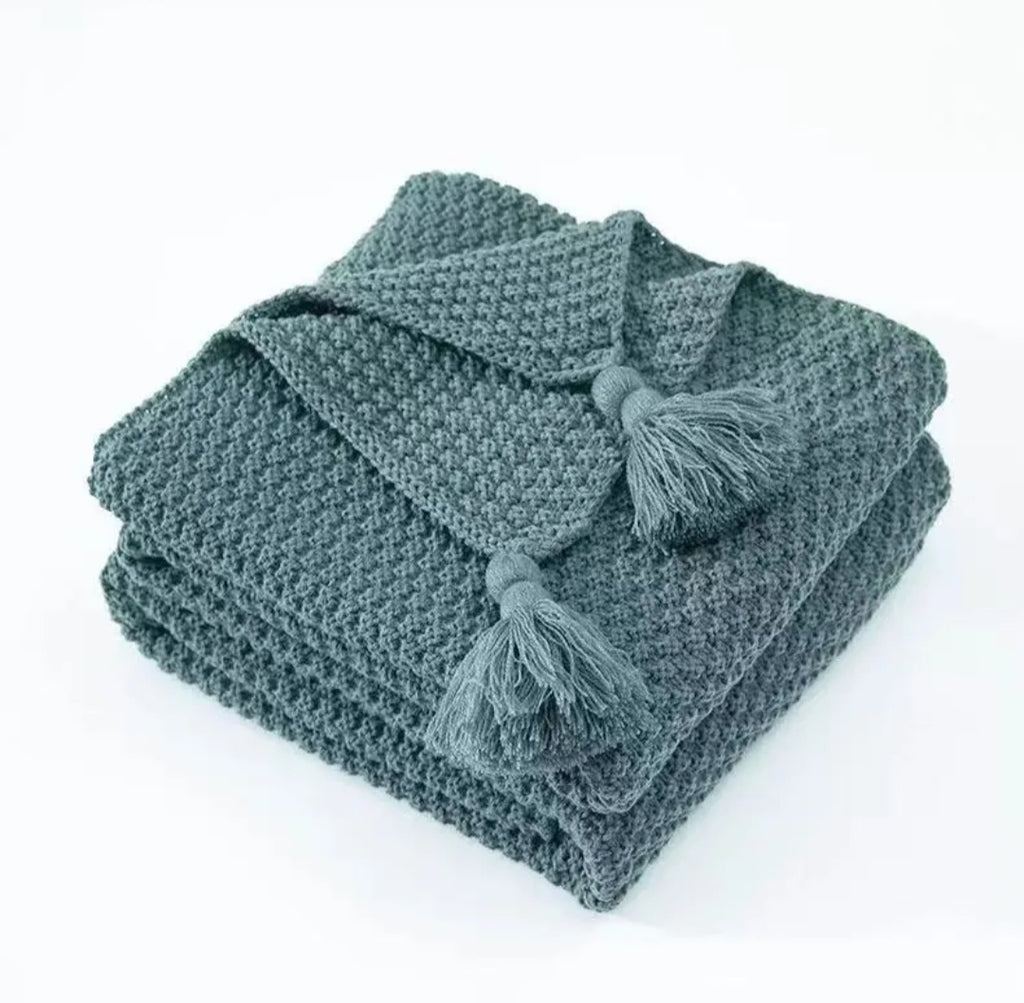 A folded textured knit throw blanket in a muted slate gray-green color, featuring two matching tassels on the corner, set against a plain white background.