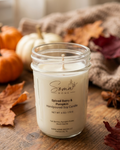 A handpoured soy candle in a clear glass mason jar by Soma Home, labeled 'Spiced Berry & Pumpkin,' set on a rustic wooden surface with autumn leaves, pumpkins, and a cozy fabric backdrop.