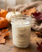 A handpoured soy candle in a clear glass mason jar by Soma Home, labeled 'Spiced Berry & Pumpkin,' set on a rustic wooden surface with autumn leaves, pumpkins, and a cozy fabric backdrop.