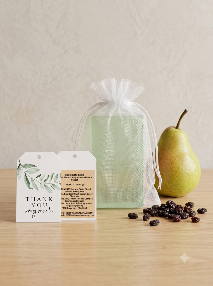 A sage green gift box wrapped in a sheer white organza bag, displayed on a light wood table next to a green pear and scattered dark raisins. Two white gift tags are placed in front of the box.