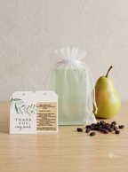 A sage green gift box wrapped in a sheer white organza bag, displayed on a light wood table next to a green pear and scattered dark raisins. Two white gift tags are placed in front of the box.