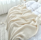 A cream-colored textured knit blanket with pom-poms draped over a bed with white bedding in a bright, softly lit bedroom.