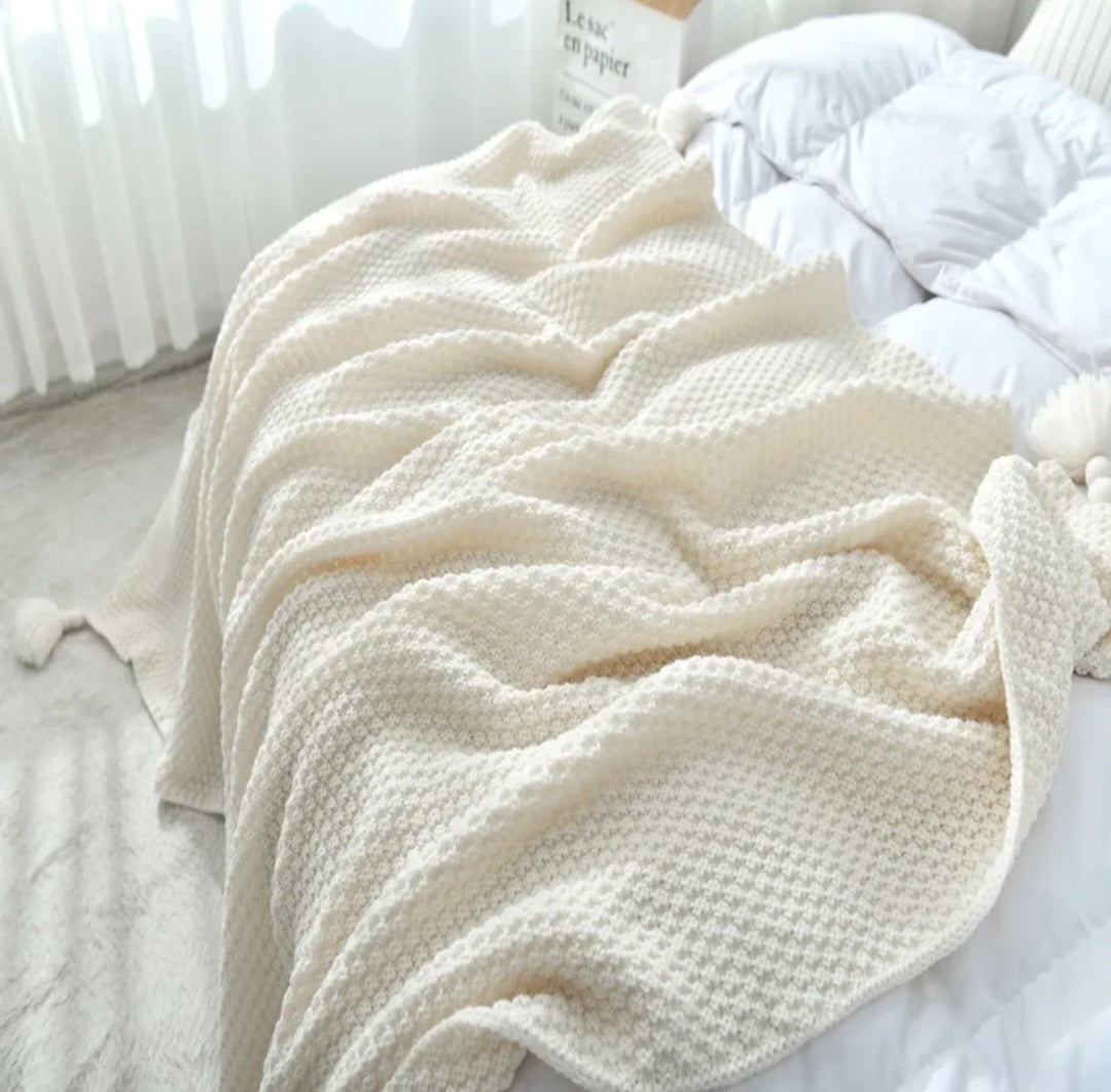 A cream-colored textured knit blanket with pom-poms draped over a bed with white bedding in a bright, softly lit bedroom.