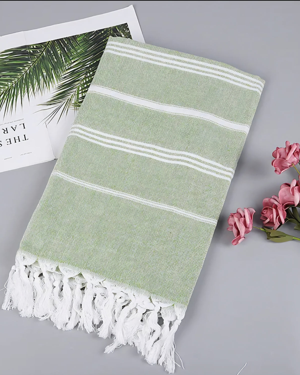 Grass green Turkish beach towel with white stripes and knotted tassels, styled with a magazine and pink roses
