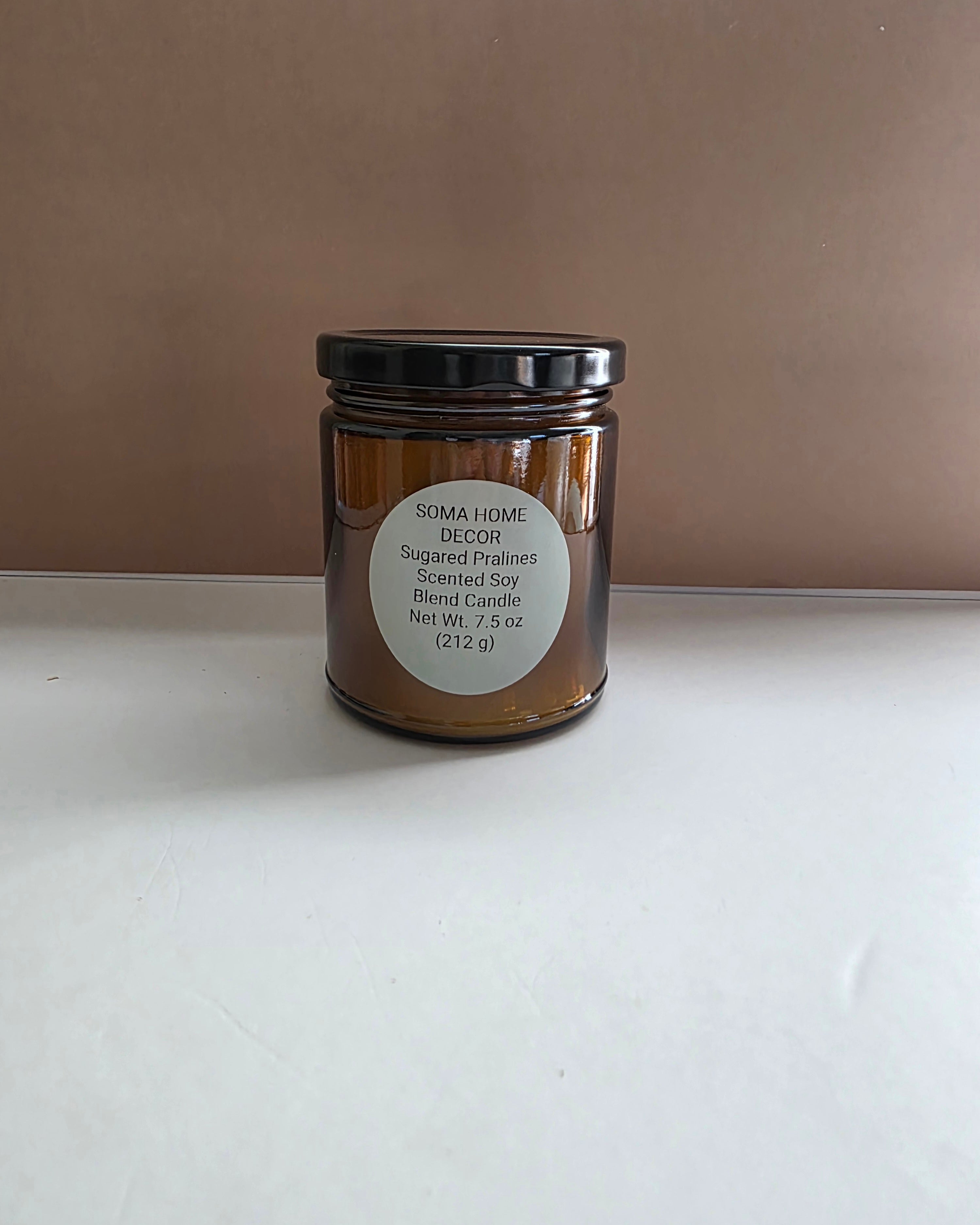 An amber glass jar candle with a black lid and a white circular label reading 'SOMA HOME DECOR Pugged Pajamers Scented Soy Blend Candle', set on a white surface against a brown background.