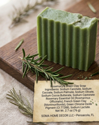A rectangular bar of green clay soap with a rustic torn edge, displayed on a wooden board with rosemary sprigs and a paper label showing ingredients and the brand SOMA HOME DECOR LLC.