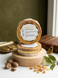A round piece of handmade soap labeled 'MONEY' stacked on a small wooden display stand, with additional soap pieces and natural ingredients like whole nuts, dried herbs, and a brass pendant arranged around it on a white surface against a dark olive background.