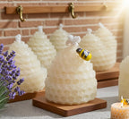 Beeswax honeycomb beehive candles displayed on wooden bases, with a small yellow and black bumblebee decoration on top of the front candle. The scene is styled with lavender flowers and a lit candle against a brick wall background.