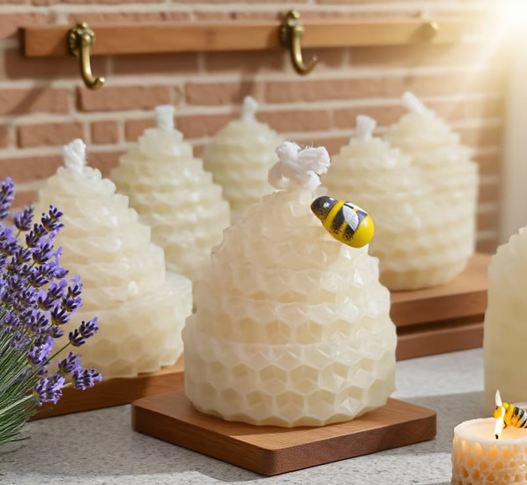 Beeswax honeycomb beehive candles displayed on wooden bases, with a small yellow and black bumblebee decoration on top of the front candle. The scene is styled with lavender flowers and a lit candle against a brick wall background.