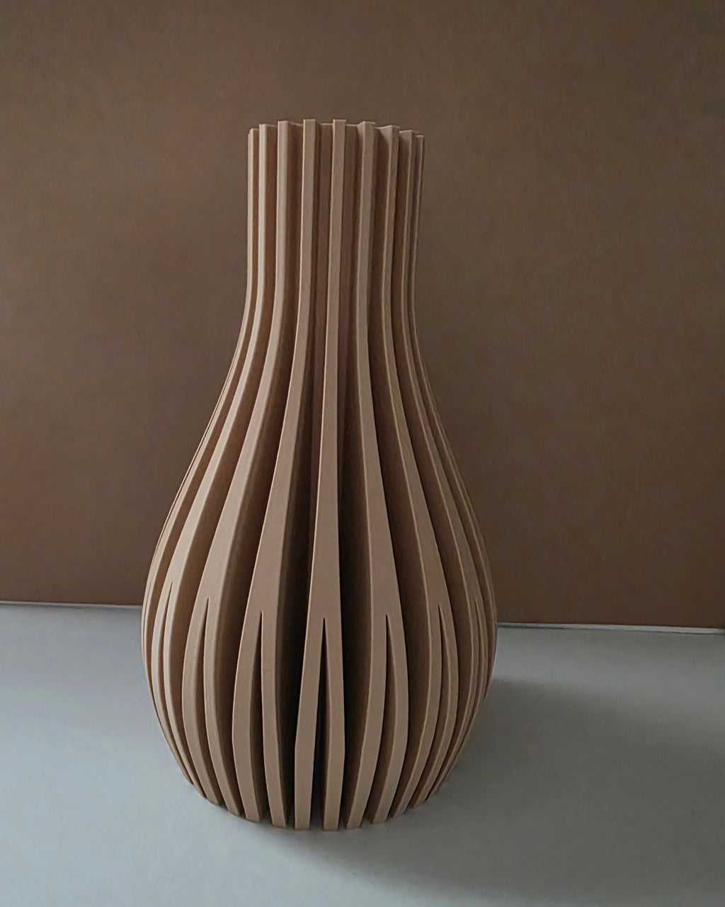A modern sculptural vase made of vertically aligned ribbed wood slats, standing on a light gray surface against a textured brown background.