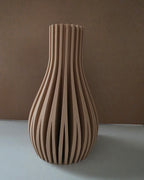 A modern sculptural vase made of vertically aligned ribbed wood slats, standing on a light gray surface against a textured brown background.