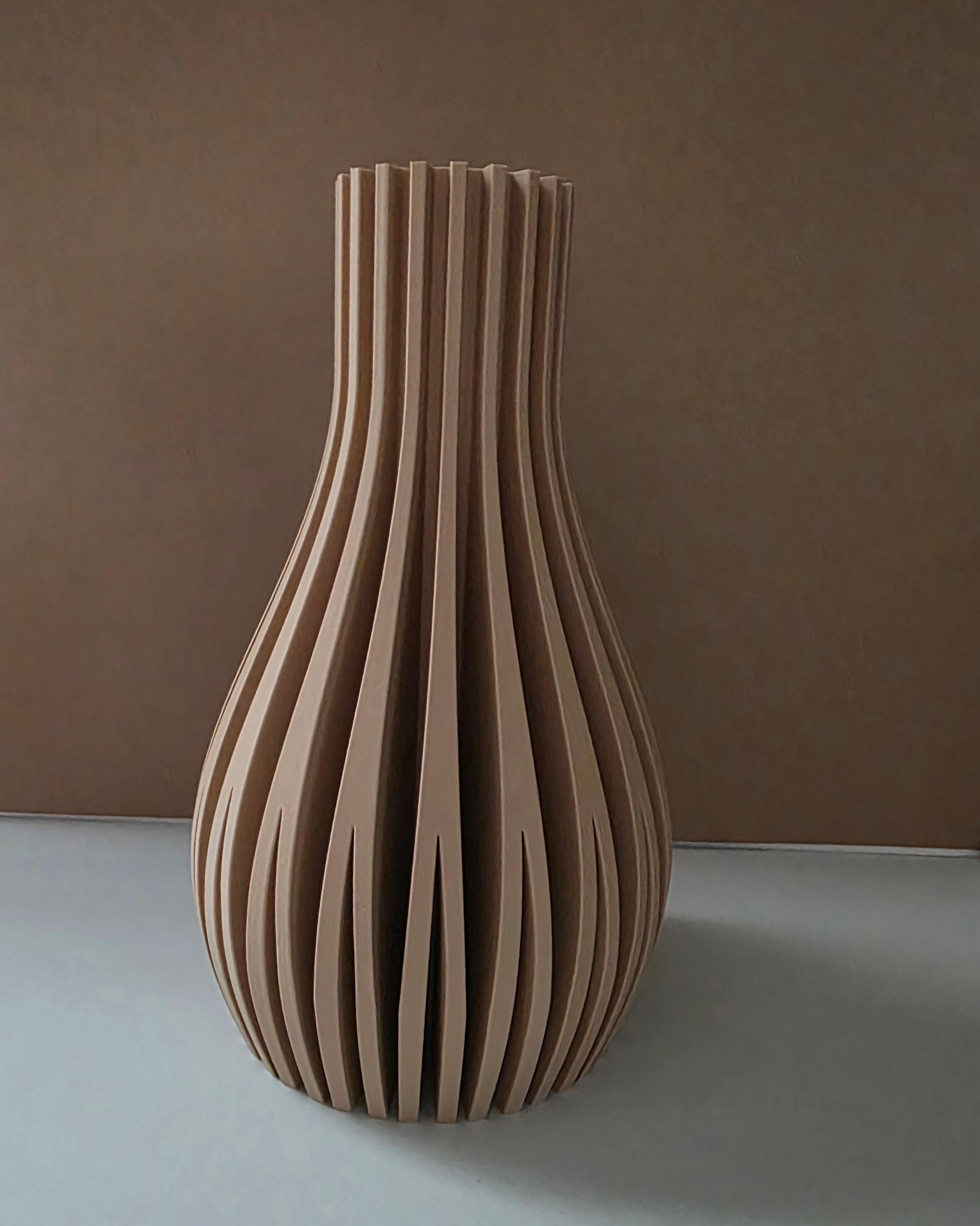 A modern sculptural vase made of vertically aligned ribbed wood slats, standing on a light gray surface against a textured brown background.