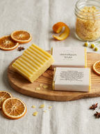 Two bars of handmade soap on a wooden board, one unwrapped showing a pale yellow bar and one wrapped in cream paper. The setup includes dried orange slices, star anise, a citrus peel, and a jar of dried botanicals on a light linen background.