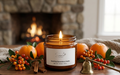 A lit amber glass jar candle labeled 'Sparking Clementine & Vanilla' by Sam's Home, styled on a wooden surface with oranges, berries, cinnamon sticks, and a brass bell in front of a blurred stone fireplace.