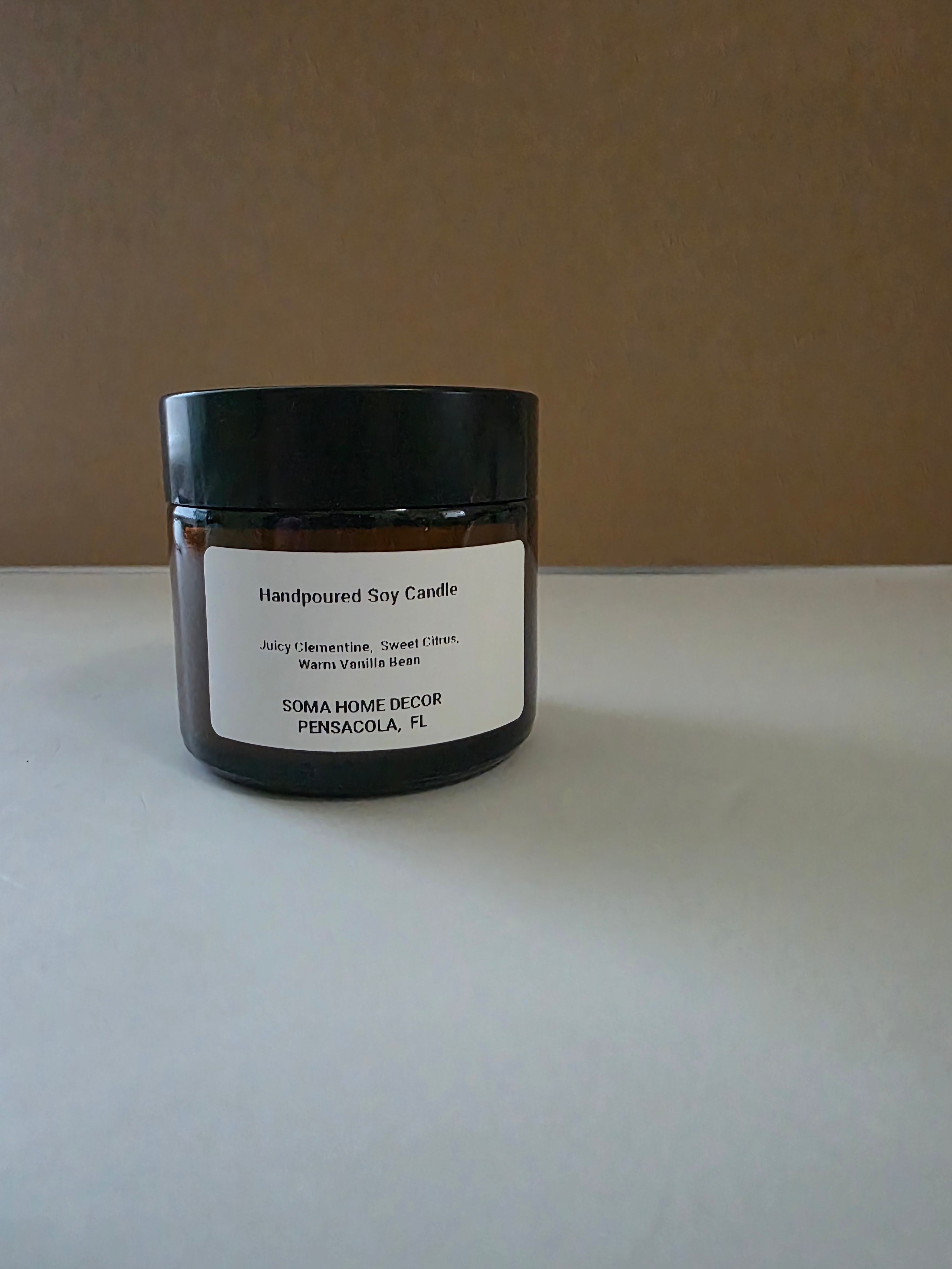 A handpoured soy candle in a dark amber glass jar with a black lid and a white label, set against a plain brown background on a light gray surface.