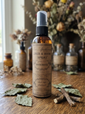 A bottle of Patchouli Linen & Room Spray by The Apothecary, set on a rustic wooden table surrounded by dried leaves and twigs, with a softly blurred background of dried flowers and glass bottles.