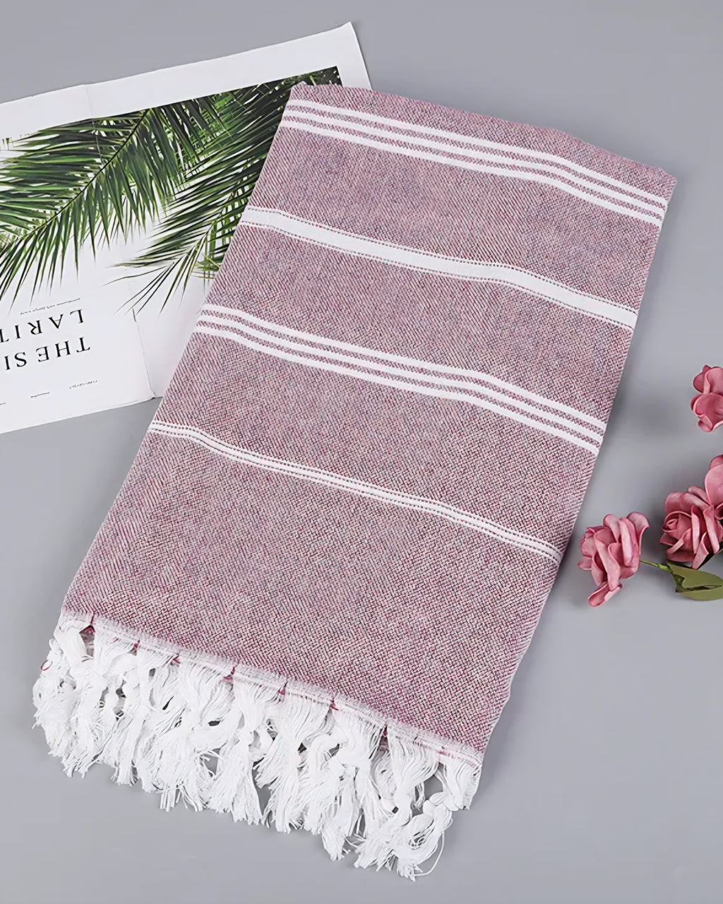 Burgundy Turkish beach towel with white stripes and knotted tassels, styled with a magazine and pink roses