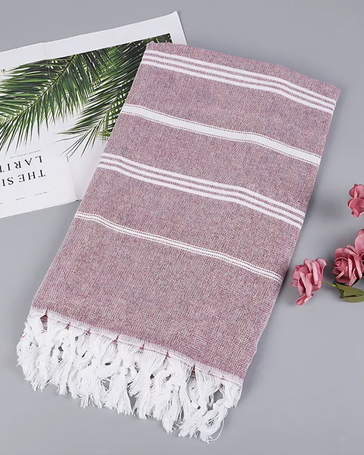 Burgundy Turkish beach towel with white stripes and knotted tassels, styled with a magazine and pink roses