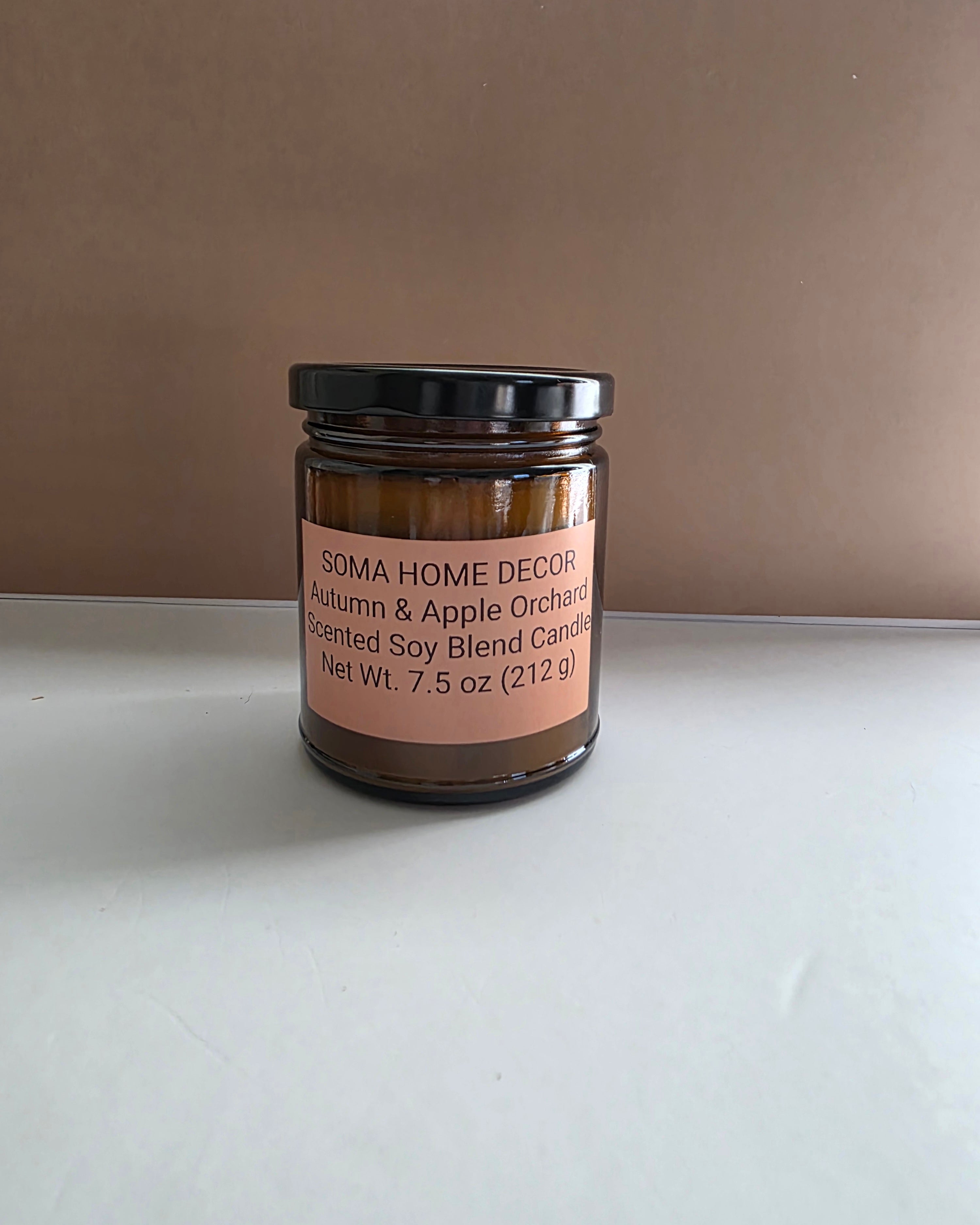An amber glass jar candle with a black lid and a peach-colored label reading 'SOMA HOME DECOR Autumn & Apple Orchard Pented Soy Blend Candle'. The candle is set on a white surface against a plain brown background.