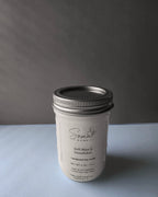 A white jar candle with a silver metal lid, labeled 'Soma' by Somal Home, placed on a light gray surface against a dark gray background.