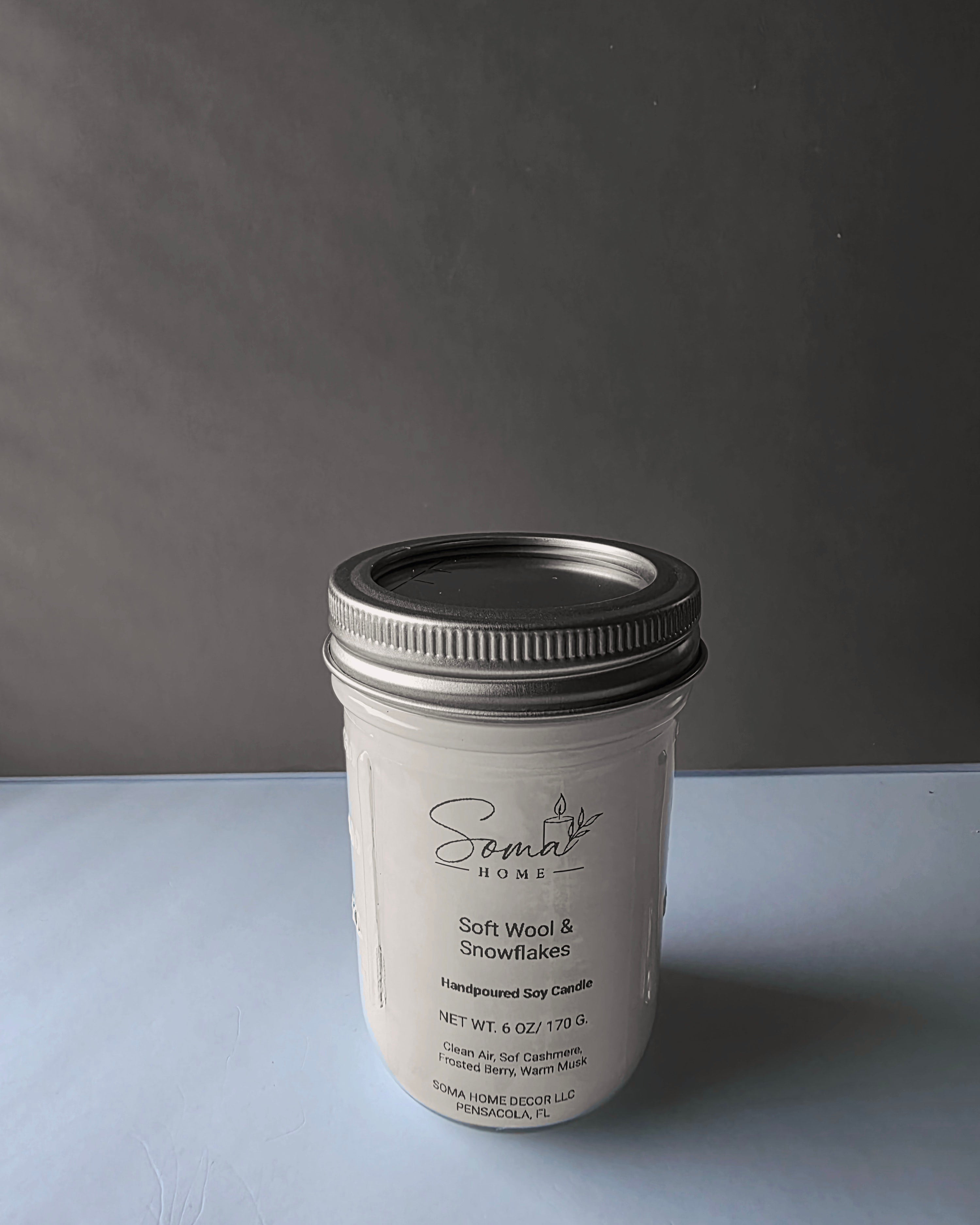 A white jar candle with a silver metal lid, labeled 'Soma' by Somal Home, placed on a light gray surface against a dark gray background.