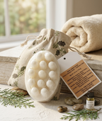 A white oval hand scrubber with raised nubs, shown with a beige botanical drawstring pouch, a product tag, a small bottle of carrier oil, stones, and greenery on a rustic tabletop. A rolled towel and soft window light are in the background.