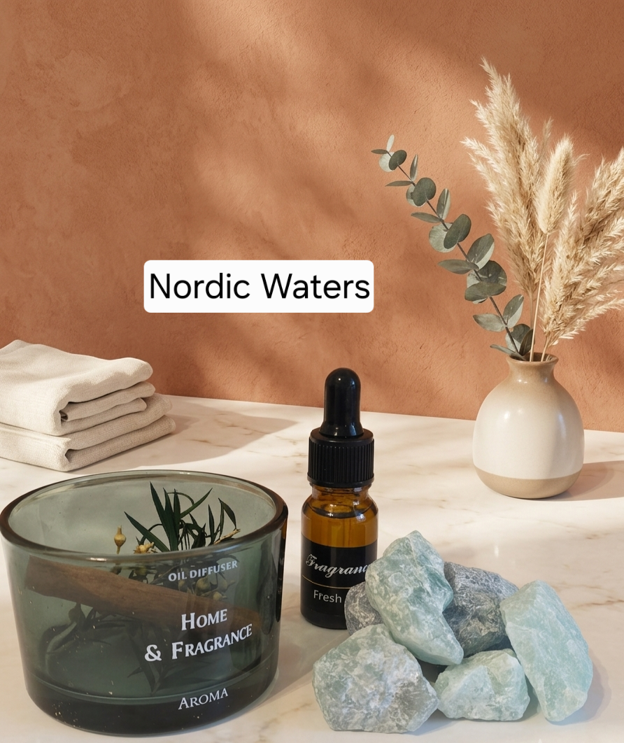 A styled home fragrance setup on a white marble surface against a warm terracotta wall. The main product appears to be a small amber dropper bottle labeled 'Nordic Waters' and 'fragrance' with a black label, alongside a dark translucent glass jar labeled 'HOME & FRAGRANCE' and pale blue crystals, with folded white towels and a ceramic vase with dried grass in the background.