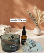 A styled home fragrance setup on a white marble surface against a warm terracotta wall. The main product appears to be a small amber dropper bottle labeled 'Nordic Waters' and 'fragrance' with a black label, alongside a dark translucent glass jar labeled 'HOME & FRAGRANCE' and pale blue crystals, with folded white towels and a ceramic vase with dried grass in the background.