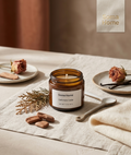 An amber glass jar candle labeled Soma Home Calmiverde Cedar is styled on a linen table runner with dried roses, a spoon, and small plates in a warm neutral setting.
