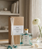 SOMA Botanical Reed Diffuser set in a softly styled home interior, featuring a clear glass bottle with dried botanicals, a kraft paper diffuser box, and white reeds, alongside a white flower in a glass vase.