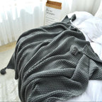 A dark gray textured knit throw blanket with pom-poms draped over a bed with white bedding, next to a book.