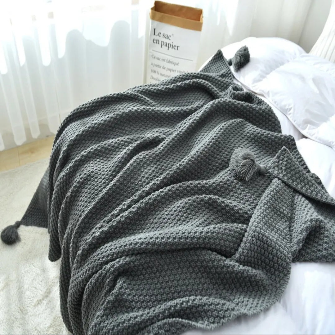 A dark gray textured knit throw blanket with pom-poms draped over a bed with white bedding, next to a book.