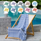 A blue and white striped Turkish cotton bath towel with fringe is draped over a wooden deck chair next to a swimming pool. Eight circular inset images at the top show the towel in different colors.