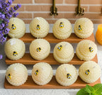 Twelve small round honeycomb-style treats arranged on two wooden trays, each topped with a small bee decoration, set on a countertop with flowers, a brick backsplash, and mint leaves around them.