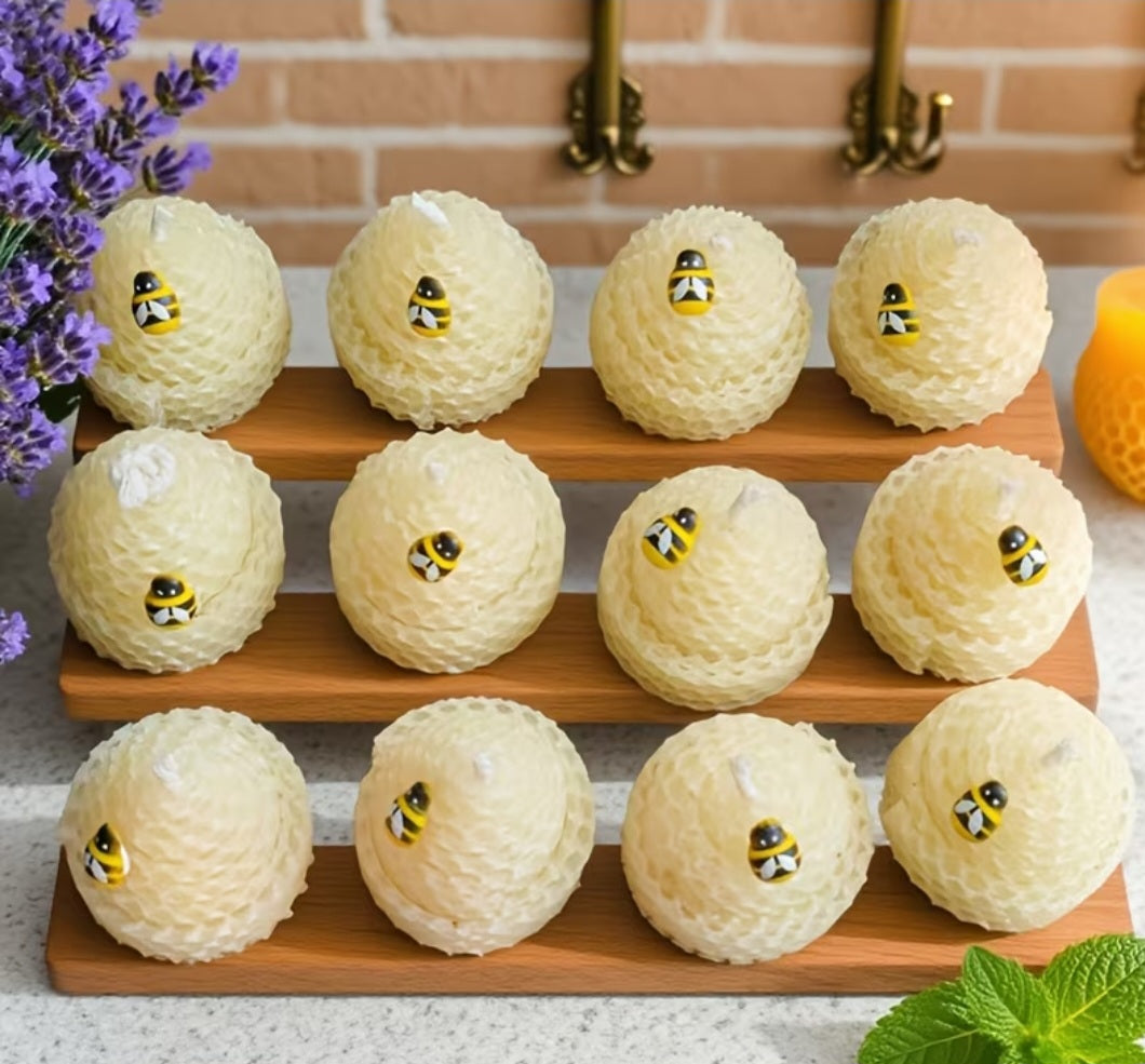 Twelve small round honeycomb-style treats arranged on two wooden trays, each topped with a small bee decoration, set on a countertop with flowers, a brick backsplash, and mint leaves around them.