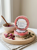 Two round bars of handmade soap on a light wood tray, styled with dried rose petals, a small bowl of loose rose petals and a wooden spoon, and dried herbs. The front soap bar is standing upright to show the label which reads 'LOVE & LUST HANDMADE SOAP ROSE PEONY & PATCHOULI MOISTURIZING, MIGHTY BUDBROKING, BALANCING'.